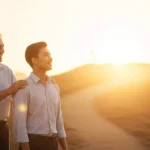 新しいキャリアの道を歩み始めた新入社員の肩に、先輩社員がそっと手を置き、明るい未来を指し示しながら励ましている心温まる風景。